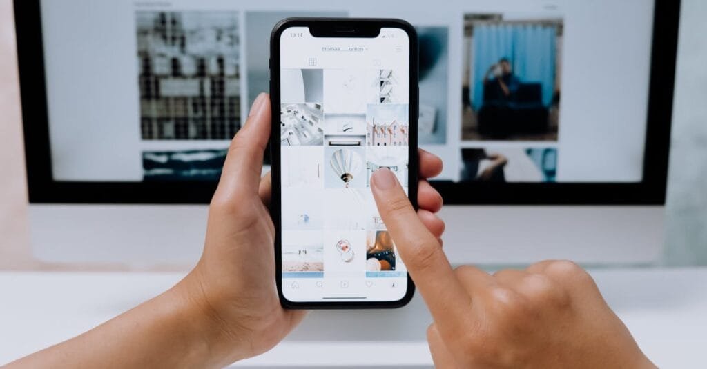 Kepentingan Website yang Mobile-Friendly untuk Perniagaan Anda 4 Hands holding a smartphone while browsing a social media app in front of a desktop computer.